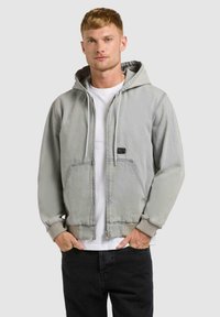 Jeune homme portant une veste à capuche gris clair zippée sur un t-shirt blanc et un pantalon noir, debout les mains dans les poches.