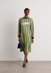 Grün gestreiftes Kleid mit langen Ärmeln, schwarzen Akzenten und einem Logo auf der Vorderseite. Das Model hält eine kleine blaue Tasche und trägt schwarze Rutschsandalen.