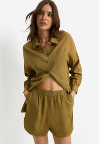 Λαδί πράσινο, ελαφρύ πουκάμισο με κουμπιά και μπροστινή τσέπη, συνδυασμένο με σορτς με ελαστική μέση. Λείο ύφασμα, casual σχέδιο, κοντή εφαρμογή.