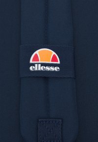 Tessuto blu navy con un'etichetta tessuta con un semicerchio arancione e rosso sopra il testo "ellesse" in bianco. Tessuto strutturato.