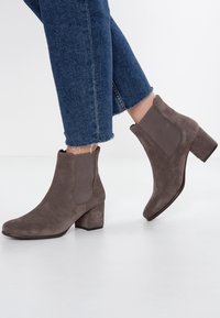 Bottines en daim marron avec un talon bloc et des panneaux latéraux élastiques. Portées avec un denim à ourlet effiloché, mettant en valeur un matériau texturé et un design épuré.