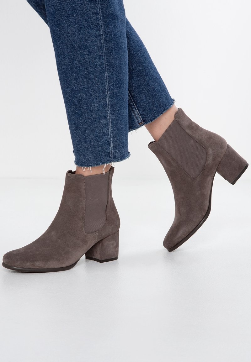 Bottines en daim marron avec un talon bloc et des panneaux latéraux élastiques. Portées avec un denim à ourlet effiloché, mettant en valeur un matériau texturé et un design épuré.