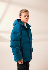 Teal polstret jakke med hætte, der har en frontlynlås og sidelommer, sat sammen med en hvid skjorte og sorte bukser.