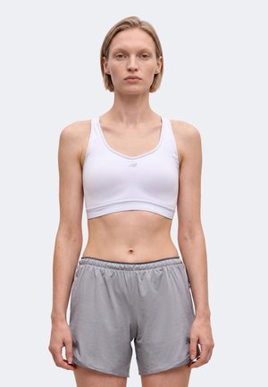 Femme aux cheveux blonds courts portant un soutien-gorge de sport blanc et un short de sport gris se tenant devant un fond clair uni.