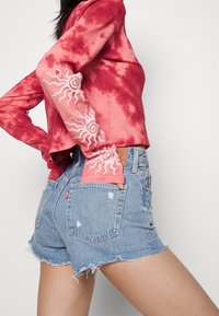 Červený batikovaný top s dlouhým rukávem a výšivkou slunce na rukávech, v kombinaci s roztrhanými džínovými šortkami s opotřebovanými detaily a koženou nášivkou.
