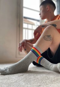 Graue knielange Socken mit einem Regenbogen gestreiften oberen Saum. Das Material wirkt weich, mit einer glatten Textur und gerippten Akzenten. Füße ruhen auf einem hellen Teppich.