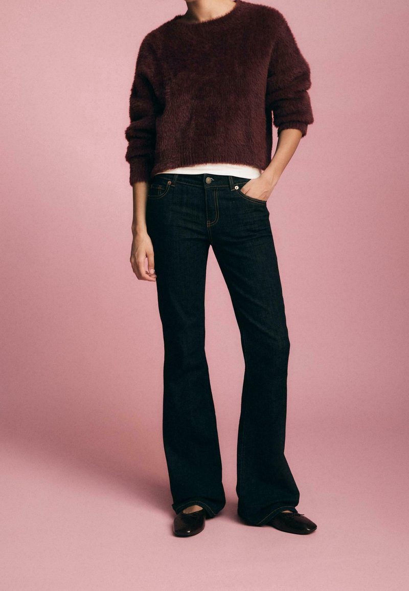 Personne portant un jean évasé foncé, un pull duveteux violet foncé par-dessus une chemise blanche et des chaussures noires, debout devant un fond rose.
