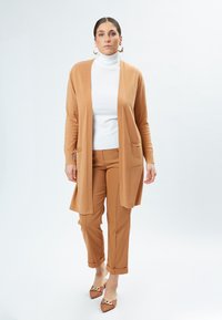 Lichtbruine lange cardigan over een witte coltrui, gecombineerd met lichtbruine broek, met een ontspannen ontwerp en twee voorzakken.