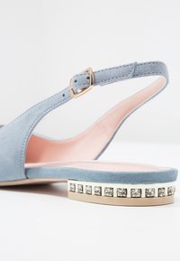 Scarpa slingback in suede blu chiaro con punta arrotondata e tacco basso piatto, decorata con borchie argentate. Interno foderato in rosa chiaro.
