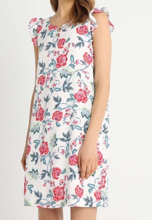 Femme portant une robe blanche longueur genou avec un imprimé floral rose et vert et des manches courtes volantées sur un fond uni.