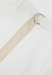 Valge voltidega kangas, millel on beež vöö, mis on läbi läbipaistva hõbedase D-rõnga pandla valgel taustal.