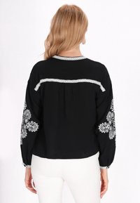 Blusa nera con delicati motivi floreali ricamati in bianco sulle maniche; presenta un collo rotondo e un delicato bordo in pizzo lungo la schiena.
