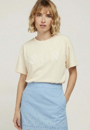 Femme blonde portant un t-shirt beige avec « SUN » en lettres florales blanches et une jupe bleu clair avec des détails œillets, main dans la poche.