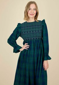Femeie cu părul blond, îmbrăcată într-o rochie din stofa cu pattern în carouri în nuanțe de verde și bleumarin, cu un decolteu brodat, stând cu o mână pe șold.