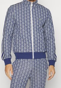 Man som bär en blå och vit geometriskt mönstrad zip-up jacka och matchande byxor med små gröna krokodil-loggor på bröstet och låret.