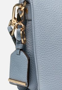 Angolo di una borsa in pelle blu chiaro con ferramenta dorata, clip per portachiavi e un'etichetta da viaggio rettangolare con un accento marrone.