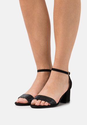 Sandales noires à bout ouvert avec un talon bloc, dotées d'une bride ornée de bijoux et d'une boucle à la cheville. Texture douce en daim et finition lisse.