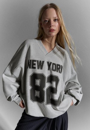 Junge Frau mit langen blonden Haaren, die ein graues übergroßes Sweatshirt mit der Aufschrift "NEW YORK 82" in schwarzen Buchstaben und eine dunkle Hose vor einem schlichten Hintergrund trägt.