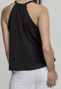 Kobieta w bezrękawniku w czarnym kolorze z wycięciem na plecach oraz w białych spodniach, pokazana z tyłu na jednolitym tle.