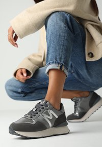 Sneaker atletici grigi con un mix di pelle liscia e suede, accenti bianchi prominenti e una robusta suola in gomma, posizionati sul piede di un modello.