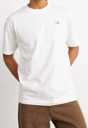 Weißes kurzärmeliges Baumwoll-T-Shirt mit rundem Ausschnitt und kleinem grauem New Balance-Logo auf der Brust; getragen zu braunen, strukturierten Hosen.