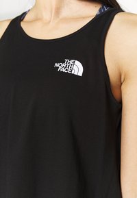 Personne portant un haut sans manches noir avec un logo blanc The North Face sur la poitrine, montrant le cou et la zone des épaules.