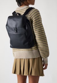 Black leather backpack featuring a flap closure, adjustable straps, and a front zip pocket. Set against a light background.