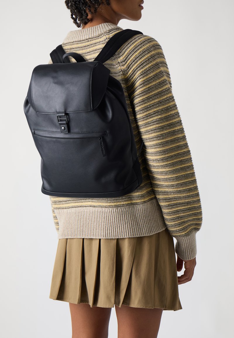 Black leather backpack featuring a flap closure, adjustable straps, and a front zip pocket. Set against a light background.
