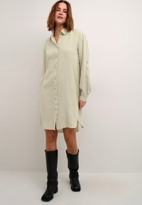 Robe chemise à boutons en vert clair avec des rayures blanches. Manches longues, fentes sur les côtés et coupe décontractée. Portée avec des bottes noires montantes.