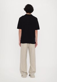 Henkilö, jolla on kiharaiset hiukset, pukeutuneena mustaan lyhythihaiseen toppiin ja väljiin beigeihin housuihin, seisoo selkä kameralle yksinkertaista valkoista taustaa vasten.