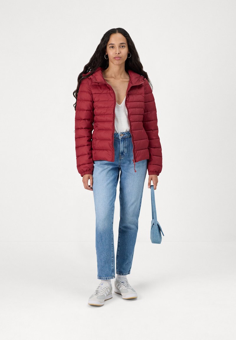 Doudoune rouge avec zip sur le devant, texture matelassée et ourlet ajustable. Associée à un jean bleu clair et des baskets grises. Petit sac à main bleu.