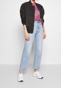 Svart bomberjacka, lila grafisk t-shirt, ljusblå vida jeans och vita sneakers med blåa och gula accenter.