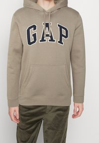 Beżowa bluza z kapturem ze sznurkiem, dużym logo "GAP" w kolorze granatowym i białym, kangurkową kieszenią z przodu, ściągaczami na rękawach oraz lekko fakturowaną tkaniną.