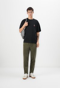 Sort bomulds t-shirt med et lille broderet design, kombineret med olivengrønne fløjelspants og hvide sneakers med brune såler. Rygsæk på skulderen.