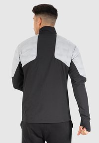 Svart jacka med grå vadderade axelpartier, hög krage, långa ärmar och en slät yta. Bakifrån visas ergonomiska designdetaljer.