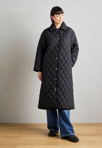 Svart quilted frakk med stor krage, med diamantmønster, knappefester og en lang, løs design. Brukt over vide bukser.