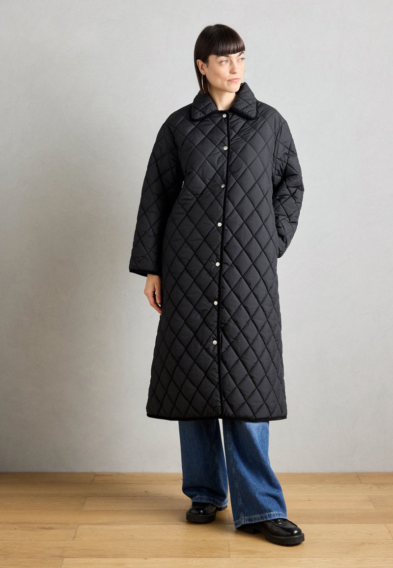 Svart quilted frakk med stor krage, med diamantmønster, knappefester og en lang, løs design. Brukt over vide bukser.