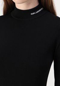 Svart langermet topp med høyhals, laget av glatt stoff, med navnet "KARL LAGERFELD" brodert i hvitt på kragen.