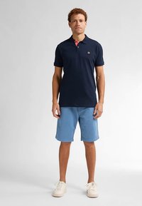 Tricou polo bleumarin cu guler roz, purtat cu pantaloni scurți bleu deschis, asortat cu adidași albi. Are un logo pe piept.