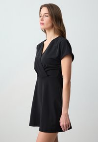 Schwarzes kurzes Kleid mit V-Ausschnitt, kurzen Ärmeln und tailliertem Schnitt. Hergestellt aus glattem, elastischem Stoff mit einem ausgestellten Rock. Einfaches, elegantes Design.