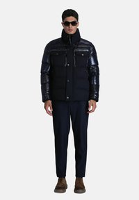 Doudoune noire avec des manches synthétiques brillantes, des poches avant et un col haut, associée à un pantalon bleu marine et des chaussures brunes.