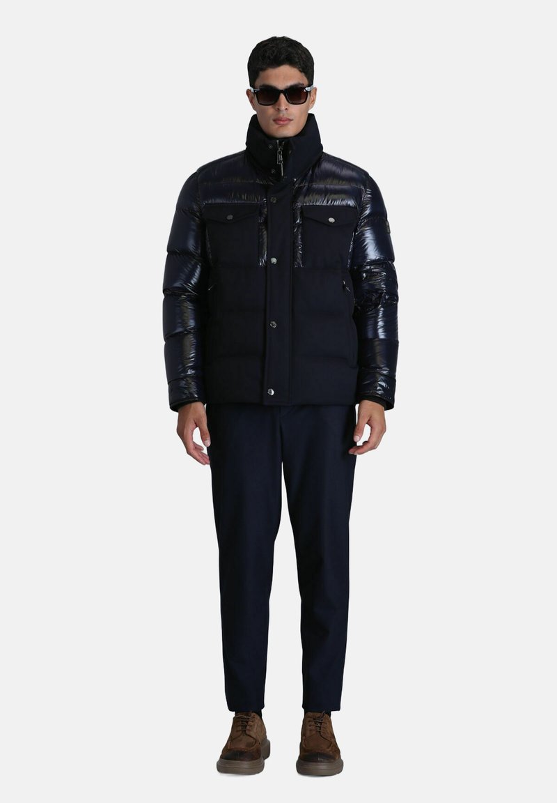 Doudoune noire avec des manches synthétiques brillantes, des poches avant et un col haut, associée à un pantalon bleu marine et des chaussures brunes.