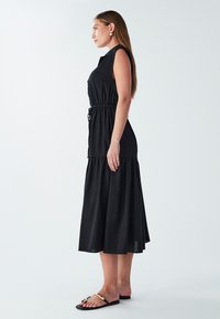 Czarna bezrękawnikowa sukienka z zaznaczoną talią, zwiewnym dołem i kieszeniami po bokach. Gładki materiał o luźnym kroju. Nosi się z płaskimi sandałami.