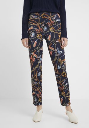 Vrouw met marineblauwe broek met een ingewikkeld patroon van kettingen, touwen en kwasten, gecombineerd met witte muiltjes met hak.