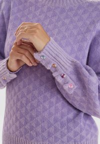 Sweter z dzianiny w kolorze lawendowym, z wzorem w romby, ściągaczami przy mankietach z kolorowymi guzikami oraz wysokim dekoltem. Miękka faktura i luźny krój.