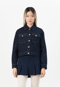 Kvinne iført en mørk denimjakke over et marineblått plisserte skjørt og hvite strømpebukser, som står foran en enkel hvit bakgrunn.
