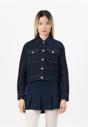 Woman wearing a dark denim jacket over a navy pleated skirt and white tights, standing against a plain white background.