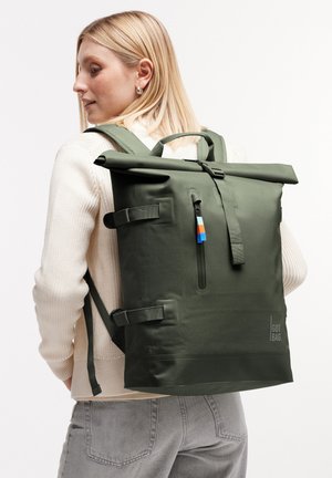 Donna bionda che indossa un maglione color crema e jeans grigi, porta uno zaino roll-top verde oliva con tasca frontale con zip e cinturini laterali.