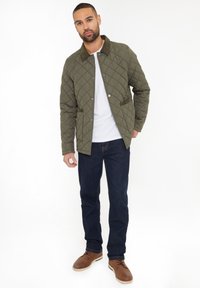 Olivenfarvet quiltet jakke med en kordfløjlskrave, to forlommer, båret over en hvid skjorte, kombineret med mørke jeans og brune sko.