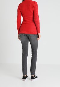 Punottu punainen neuletakki, jossa on teksturoitu kuosi ja istuva vyötärö, yhdistettynä harmaisiin kapeisiin farkkuihin ja mustiin kangasliukuisiin kenkiin.